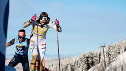 Två längdskidåkare tävlar i en uppförsbacke i vinterlandskap, med snötäckta träd och klarblå himmel i bakgrunden.