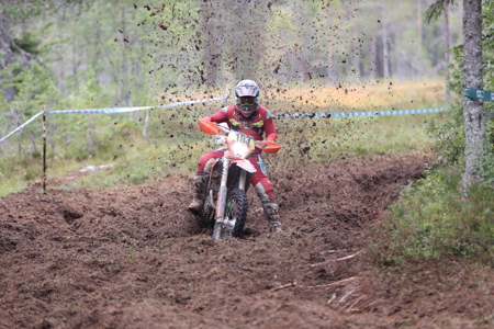 Motocrossförare i röd skyddsutrustning kör en orange motorcykel genom en lerig skogsbana, med lera som sprutar upp runt föraren.