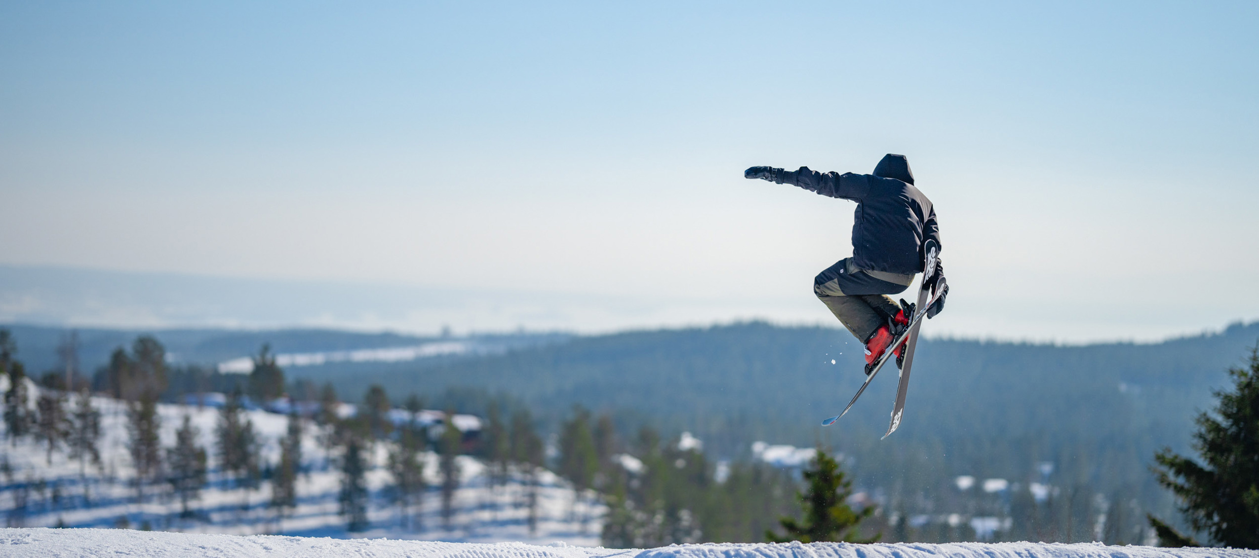 Skidåkare gör ett hopp i en snowpark med snölandskap och skog i bakgrunden.